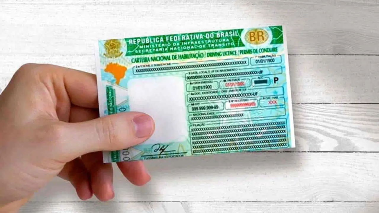 A imagem mostra uma pessoa segurando a Carteira Nacional de Habilitação (CNH) do Brasil, com o fundo em um tom neutro e madeira aparente. A CNH é um documento importante para motoristas brasileiros, que atesta a sua aptidão para dirigir, sendo emitido pelo Departamento de Trânsito (DETRAN).
